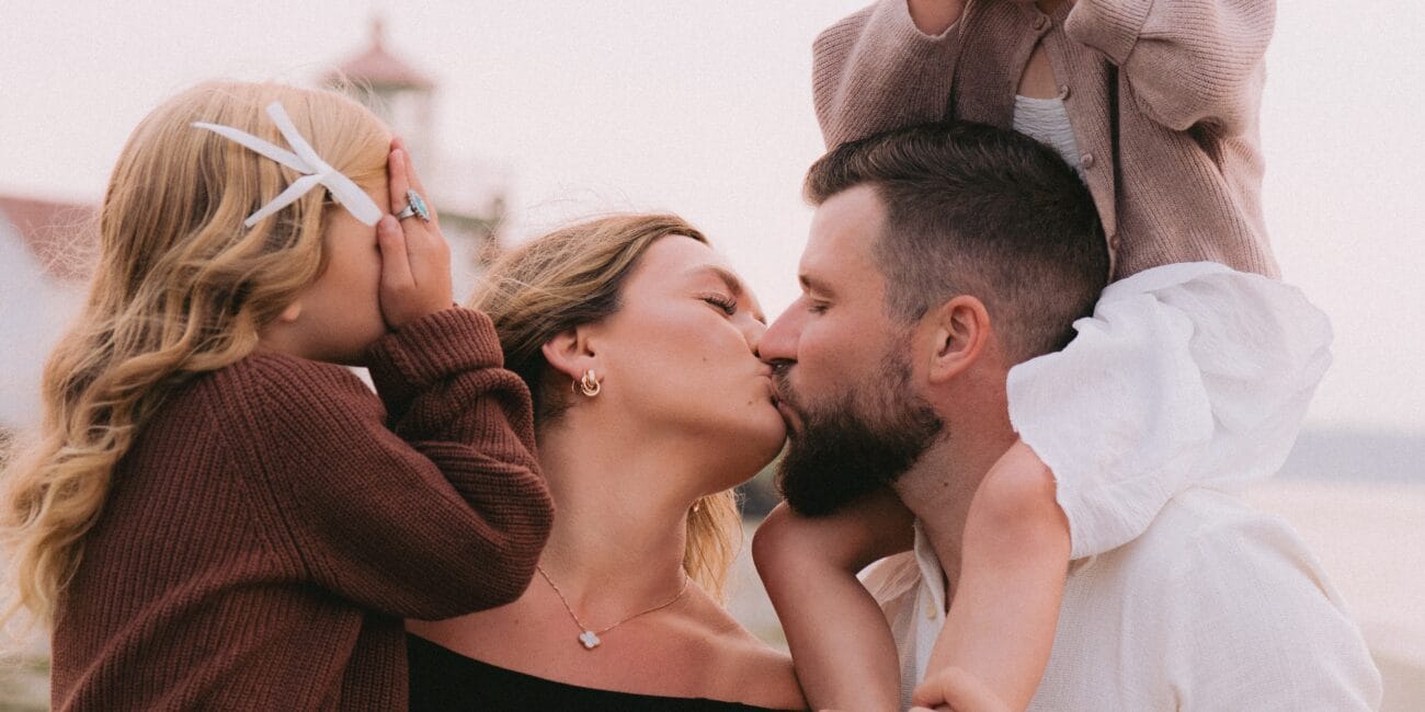A family of four on the beach, with parents kissing and two children covering their eyes.