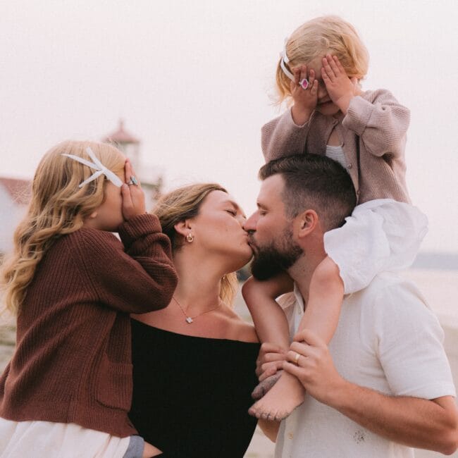 A family of four on the beach, with parents kissing and two children covering their eyes.