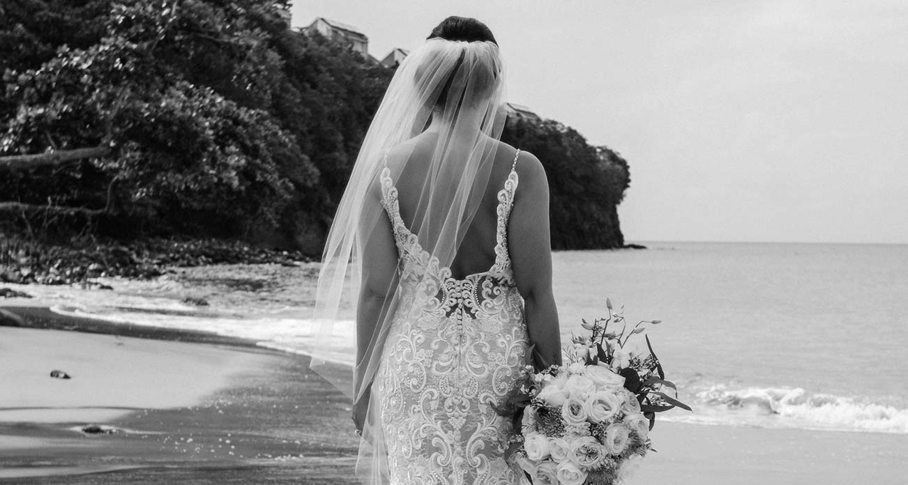 Bride in elegant wedding gown with bouquet on a beach shoreline.