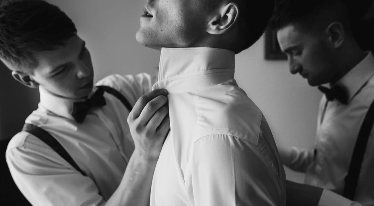 Two men in formal attire adjusting each other's shirts and bowties.