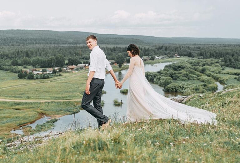 Bride and groom holding hands walking in a grassy field with a river and trees in the background.