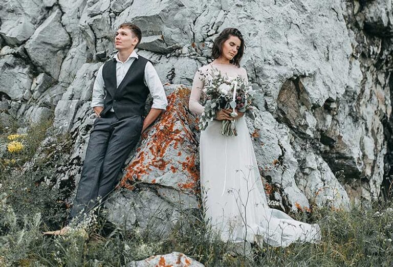 Bride in white dress holding bouquet, groom in vest standing against rocky cliff
