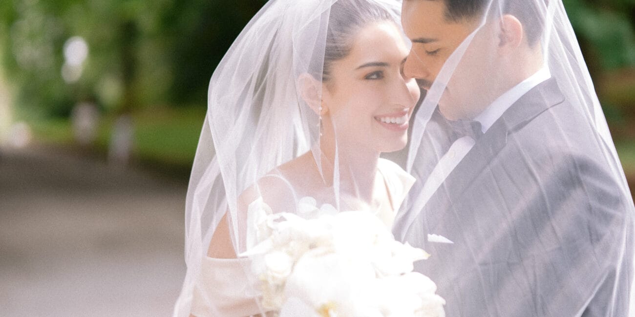 Bride and groom smiling under veil with white orchid bouquet in an outdoor setting.