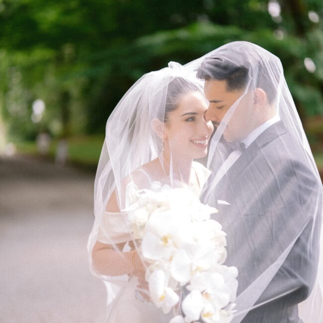 Bride and groom smiling under veil with white orchid bouquet in an outdoor setting.