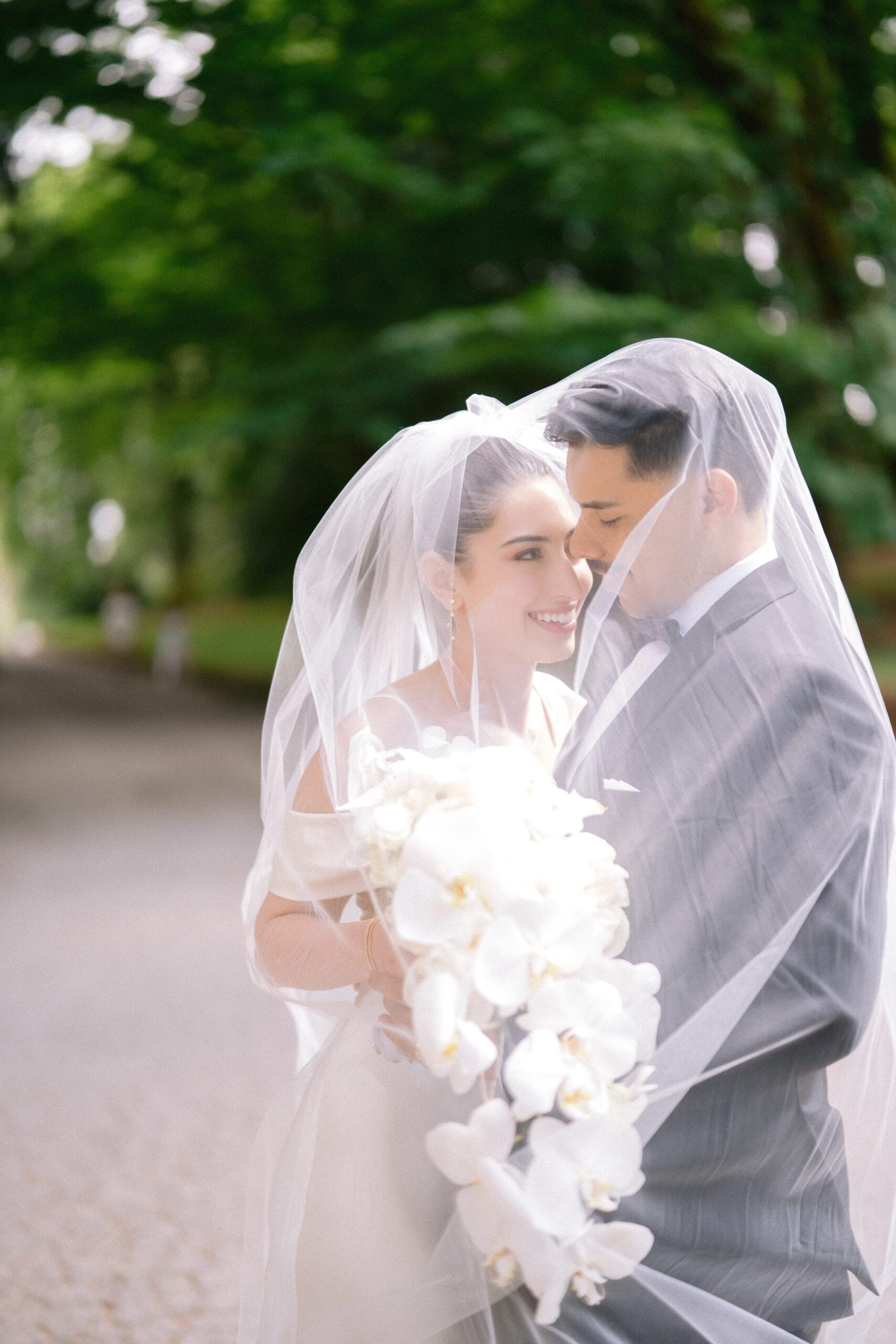 Bride and groom smiling under veil with white orchid bouquet in an outdoor setting.
