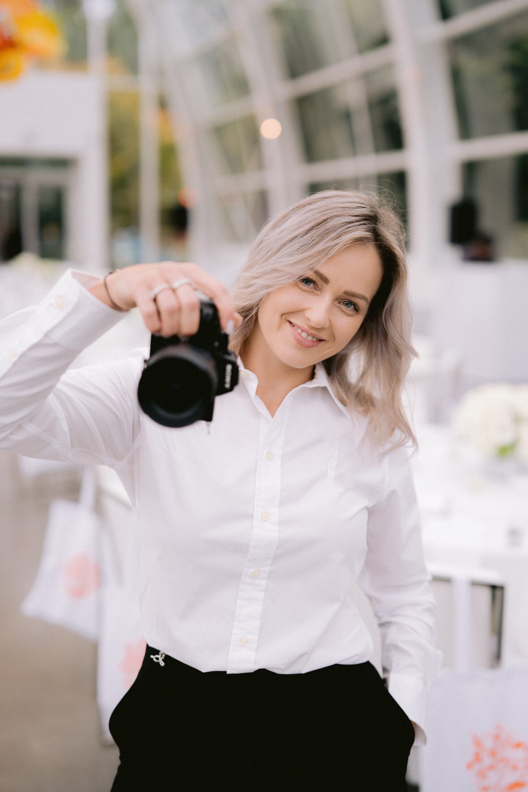 Smiling woman in white shirt holding a camera indoors.